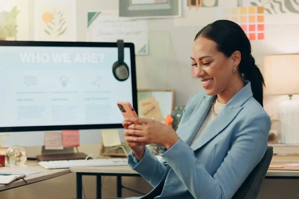 A smiling businesswoman using a smartphone in an office with a computer display in the background