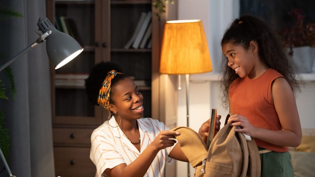 A smiling woman helps a young girl pack a backpack